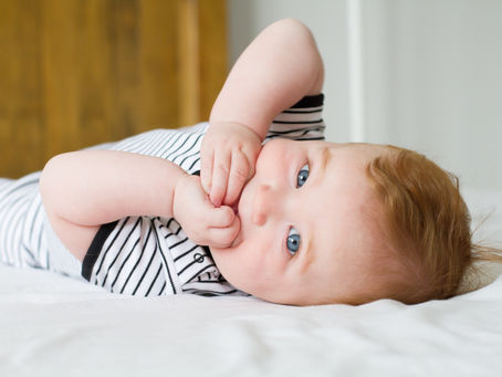 baby chewing fingers whilst laying on a bed looking at the camera