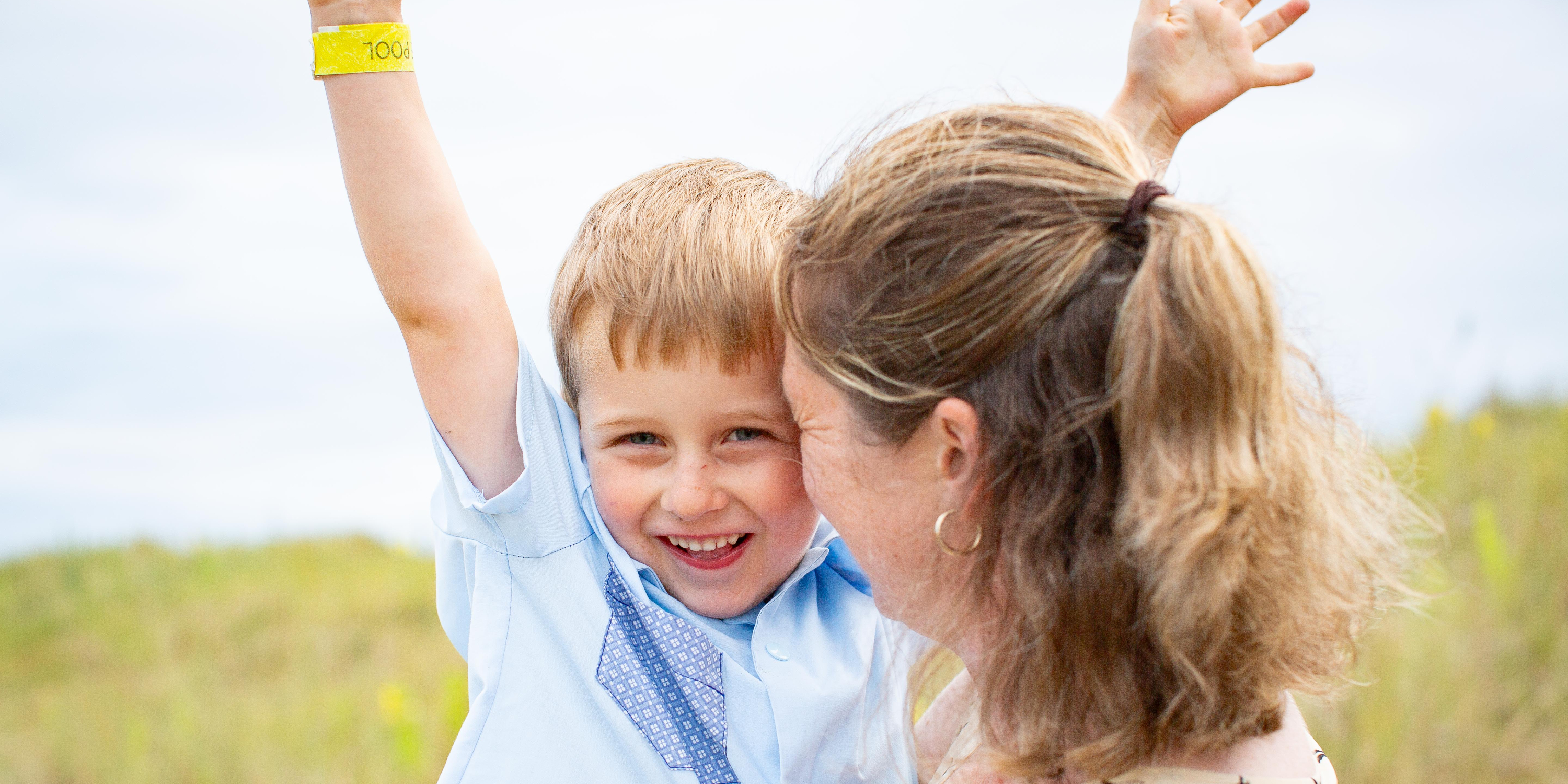Smiling boy in a blue shirt raises both arms while a woman hugs him outdoors in a sunny field, joyful celebration