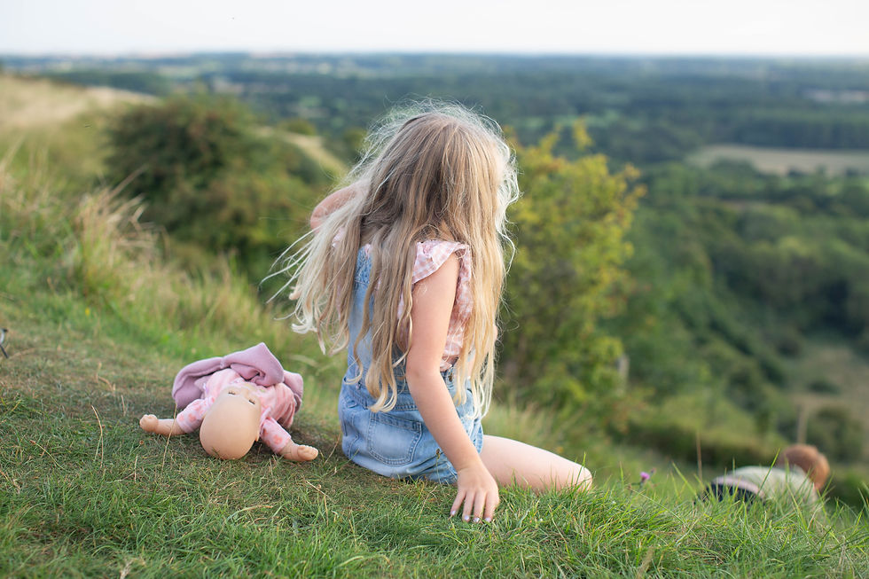 Girl with long hair sits on grassy hill, beside her a doll in pink. Scenic green landscape stretches out in the background. Peaceful.