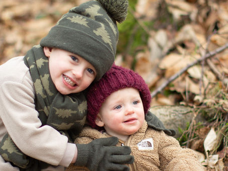 brother and sister wearing hats and scarves sitting in the autumn leaves