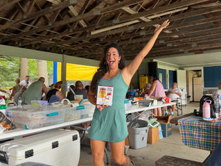 Smiling woman in teal dress poses under a pavilion, holding a Slap Chop Press box while people mingle at tables behind her