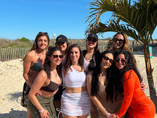 Eight women smile and pose on a sunny beach by a palm tree and sand dunes under a clear blue sky.