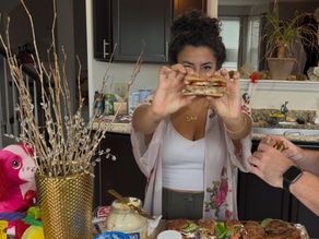 Woman in a kitchen holds up a stacked sandwich toward the camera, with festive decor and more sandwiches on the counter.