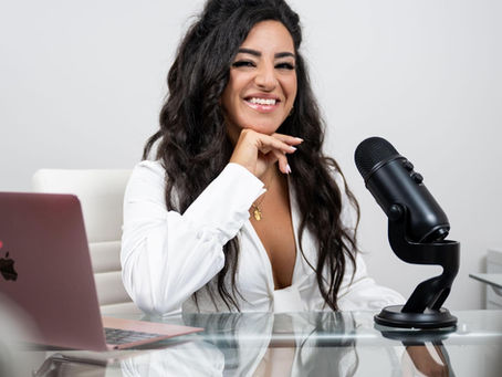 Woman with long hair smiles at a desk with a black microphone and pink laptop on a glass surface, wearing a white shirt in an office setting. Shanon Safi, host of the Fuel the Fire Podcast