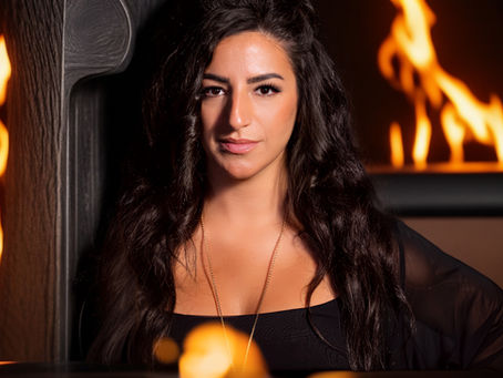 Woman with long dark hair in black top, standing near flames in background. Warm glow creates a dramatic and intense atmosphere.