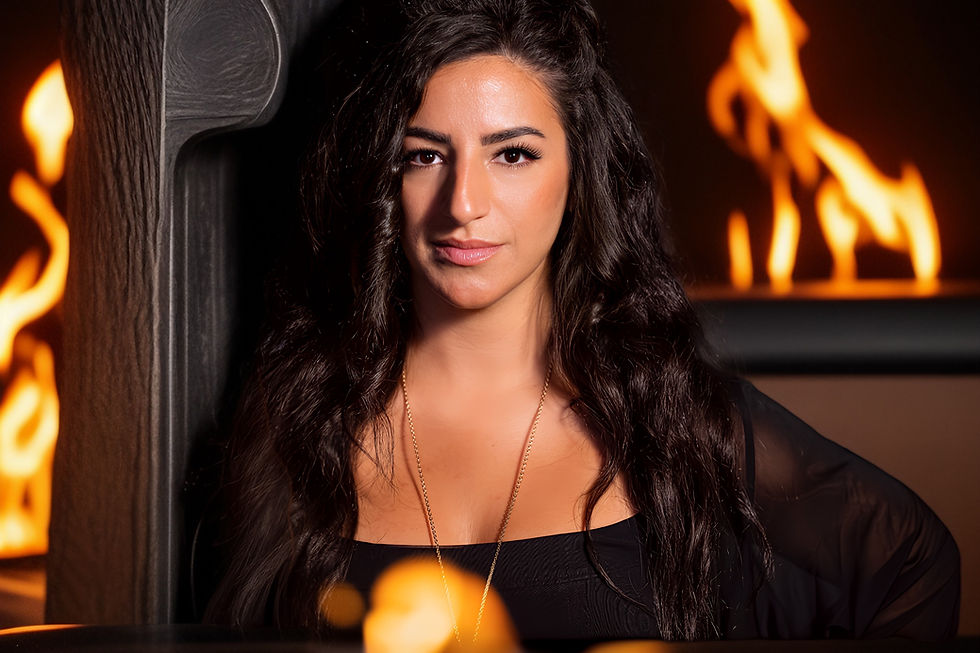 Woman with long dark hair in black top, standing near flames in background. Warm glow creates a dramatic and intense atmosphere.