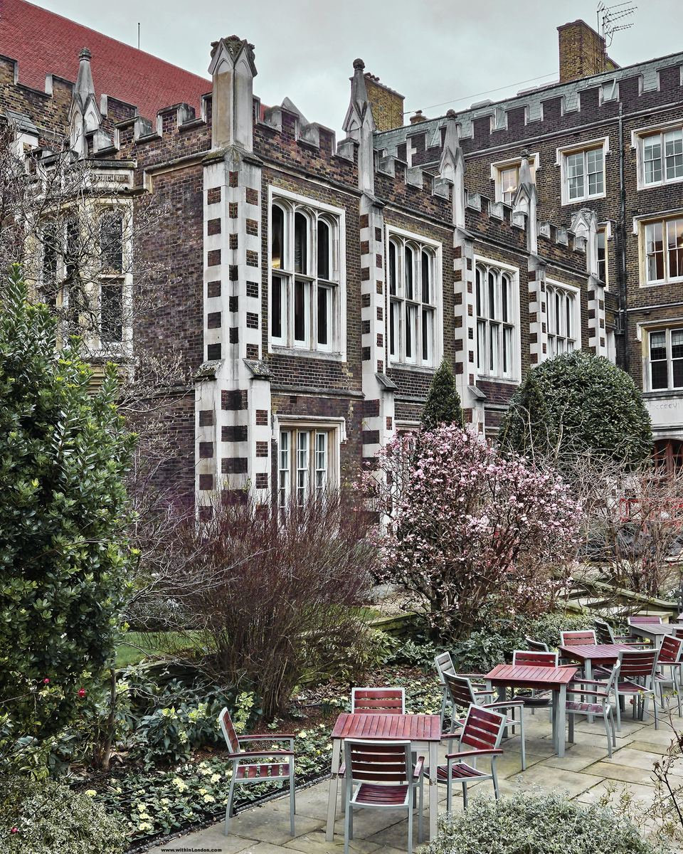 Tudor-style brick and stone building with a crenellated roofline, a magnolia tree and garden tables in front