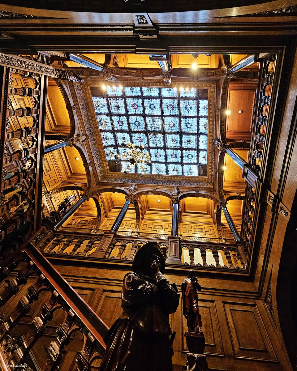 View up a carved wooden staircase towards a coloured glass skylight, with a carved wooden figure on the newel post