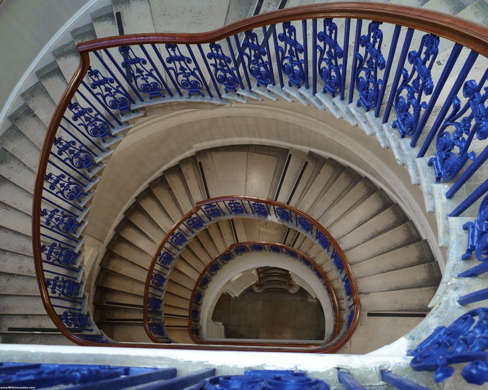 Looking straight down the oval stone staircase at the Courtauld, blue and gold ironwork spiralling to the floor below
