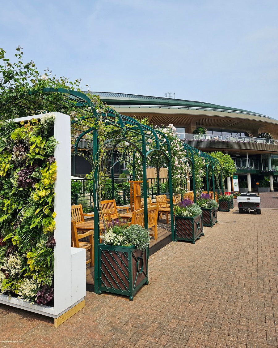Paved garden with green living walls, a pergola and planters, and the curve of a stadium behind