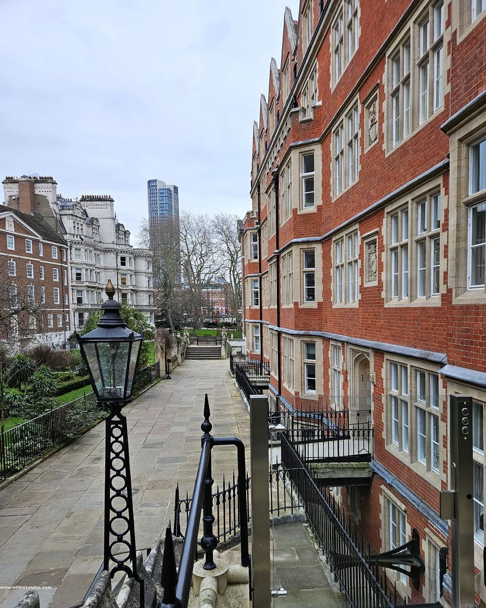 Paved walk beside red-brick chambers with a black lamp post, lawns below and a tall tower in the distance