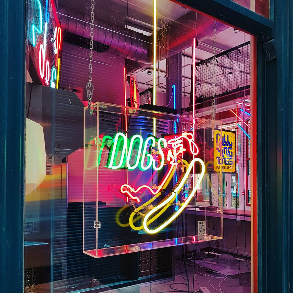 An illuminated neon 'Hot Dogs' sign shaped like a hot dog, on display in a shop window.