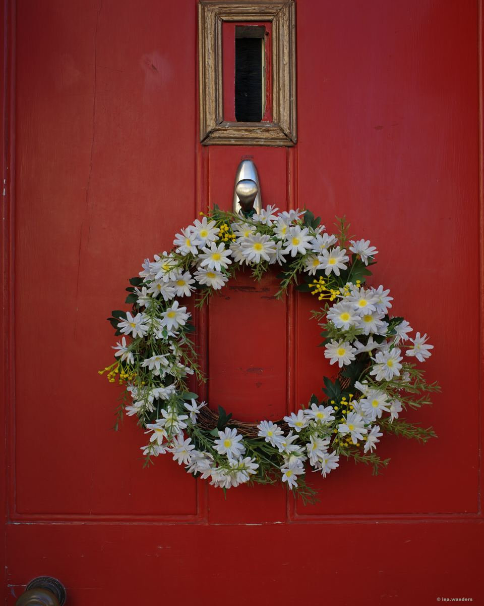 Red front door with a wreath of white daisies and a small square window