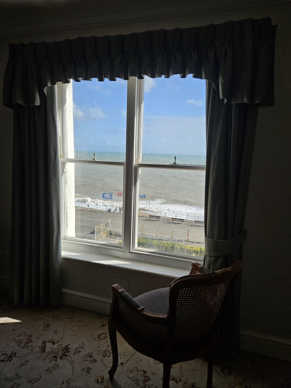 Hotel room window framed by heavy curtains, a wooden chair in front and a grey sea outside