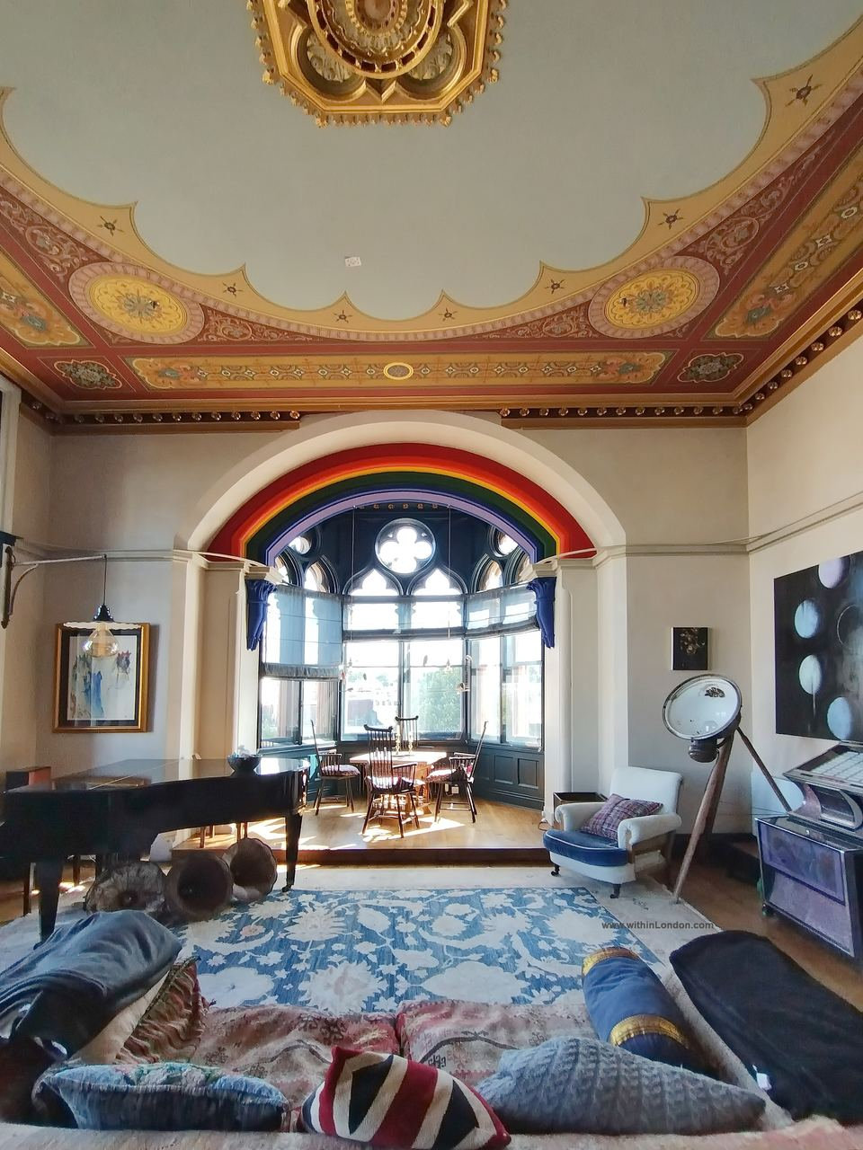 Low sofa piled with cushions on a blue patterned rug, looking across the drawing room to the rainbow-painted arch and bay window