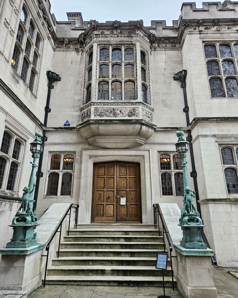 Pale stone neo-Gothic front with an oriel window and a wooden door, steps flanked by two green bronze lamp standards with small figures