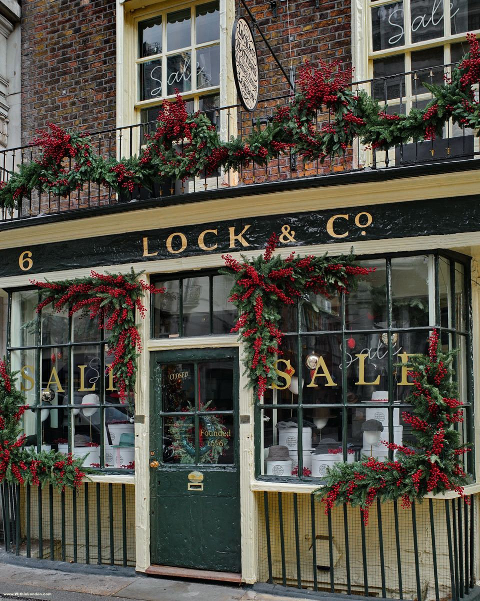 The green shopfront of Lock & Co Hatters on St James's Street, with garlands, red flowers and a sale sign in the bow windows
