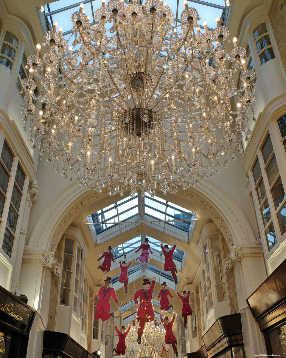 A large crystal chandelier under the glass roof of Burlington Arcade, with small figures in red hanging below it