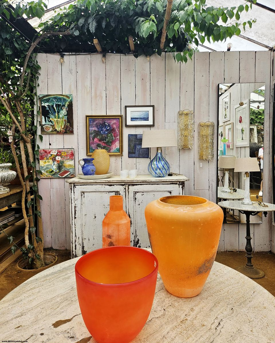 Orange glass vases on a round stone table in front of a whitewashed wooden wall hung with paintings