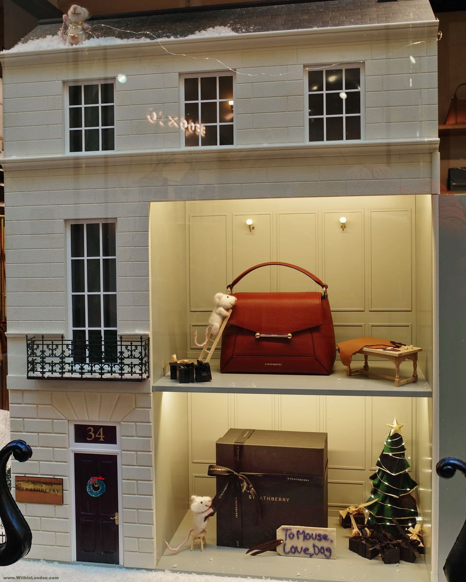 A miniature white townhouse numbered 34 in a shop window, with a red handbag on the upper floor, a decorated tree downstairs and small white mice climbing a ladder