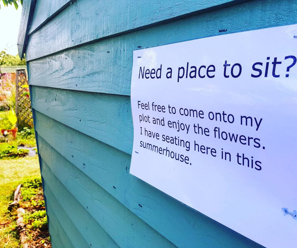 Handwritten notice on a pale blue allotment shed inviting visitors onto the plot to sit in the summerhouse and enjoy the flowers