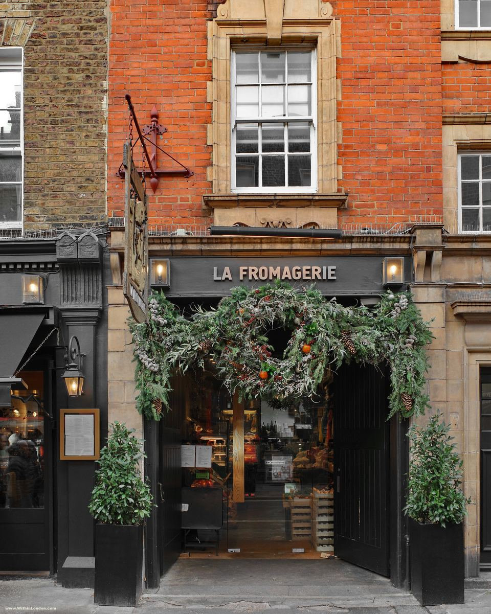 A thick arch of green foliage framing the door of La Fromagerie on Moxon Street, between two potted trees