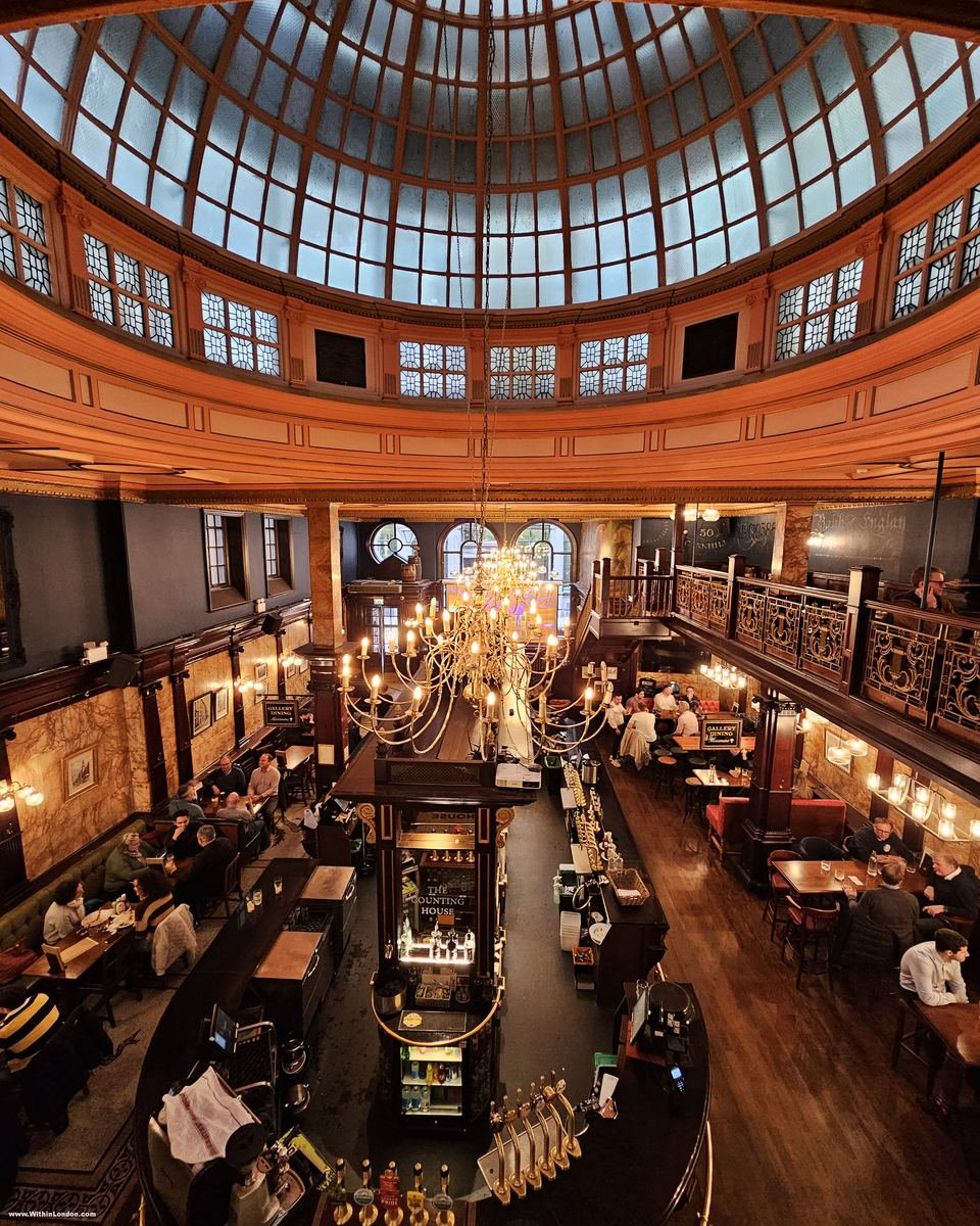 Glass dome spanning a Victorian banking hall, with an oval bar and crowded tables below, seen from a gallery balustrade