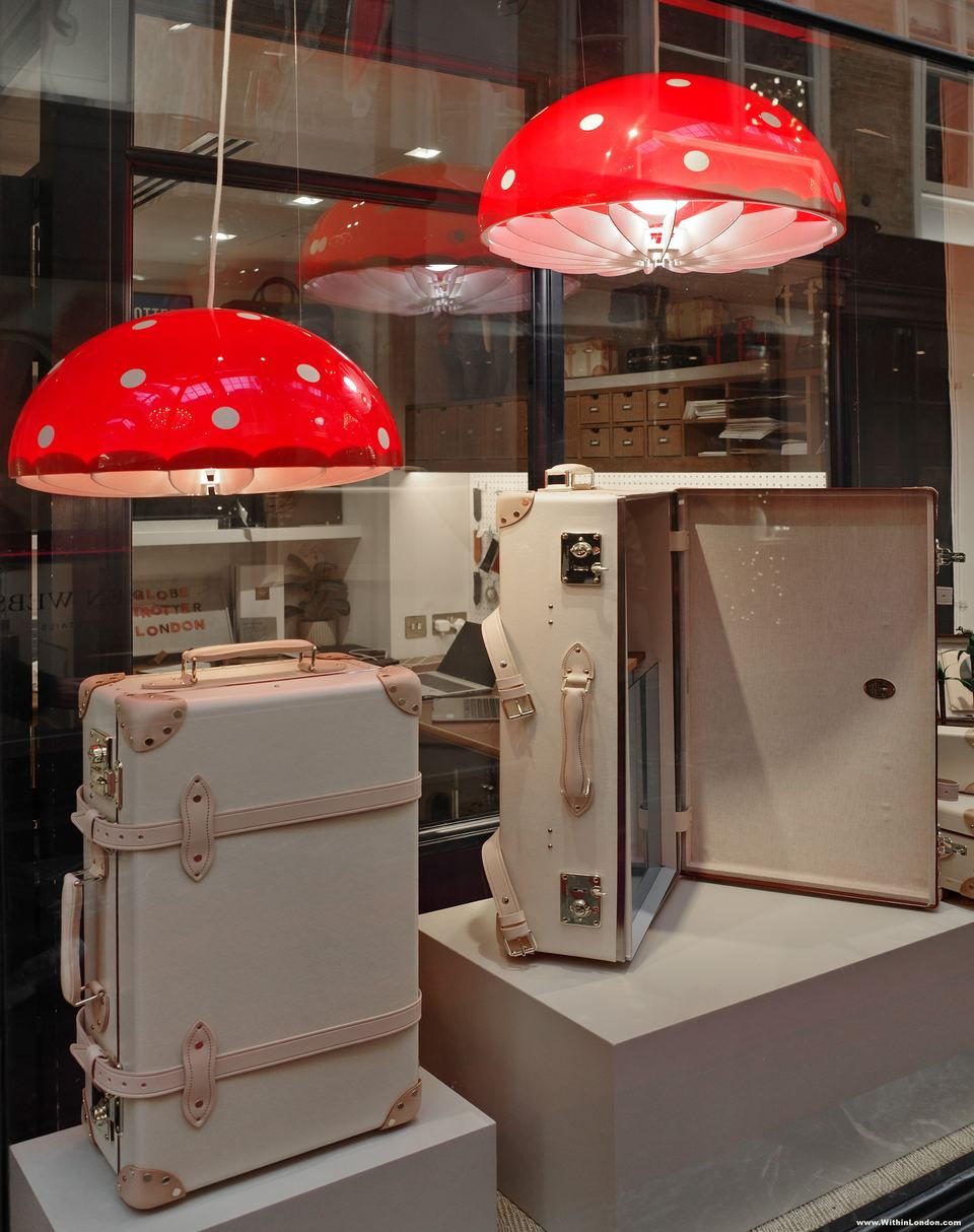 Two red toadstool lamps with white spots hanging over cream suitcases with pink leather trim in a luggage shop window