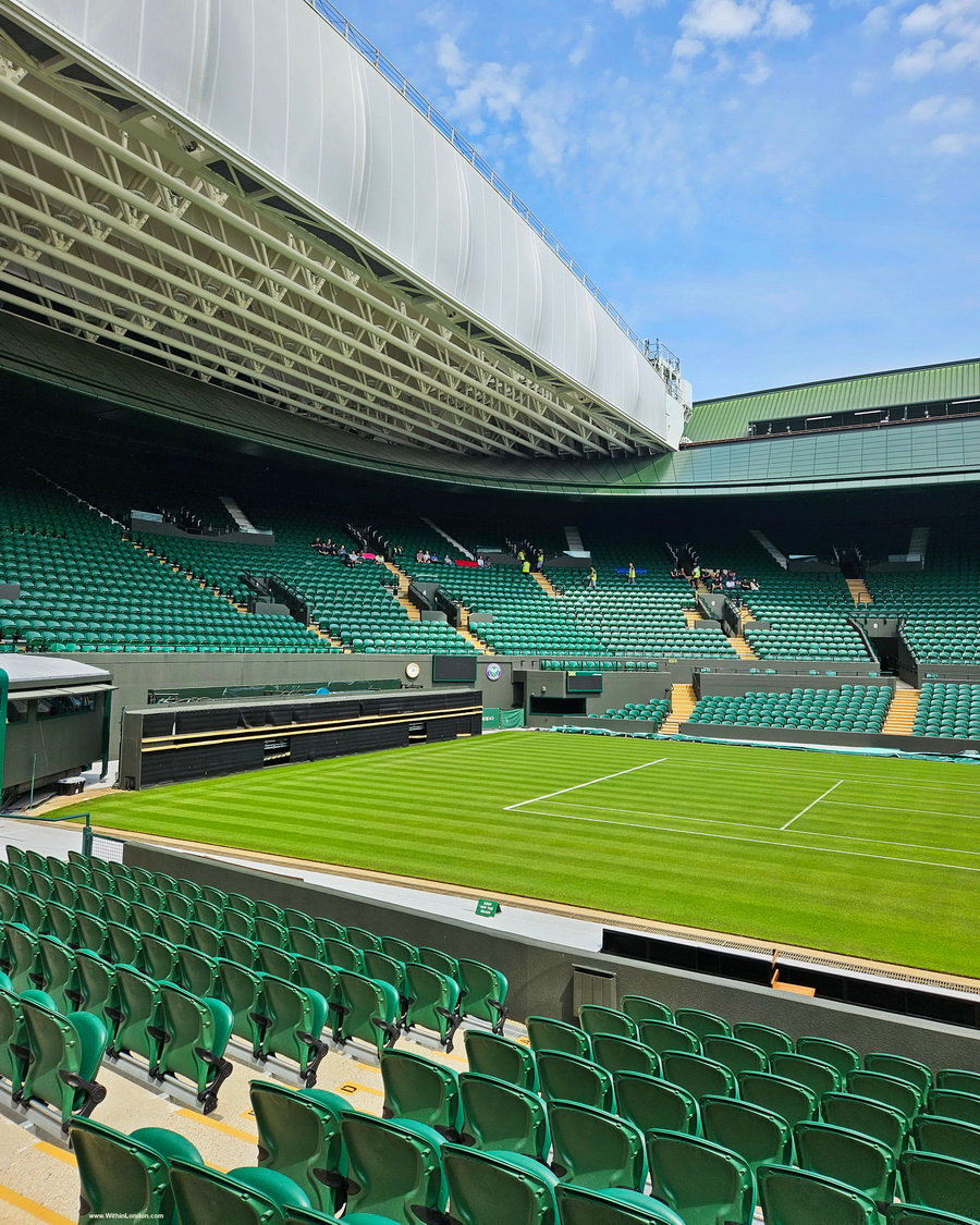 Empty green seats of No. 1 Court at Wimbledon under the curved edge of its roof, with the grass court and blue sky