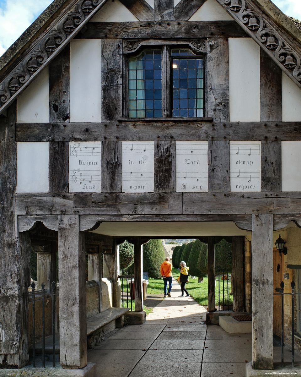 Timber-framed lychgate at St Mary's Church with music-notation panels above the arch and two people walking through to the yews