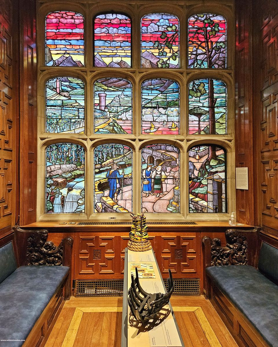 Large stained-glass window of landscape scenes above carved panelling and benches, with a display case in front