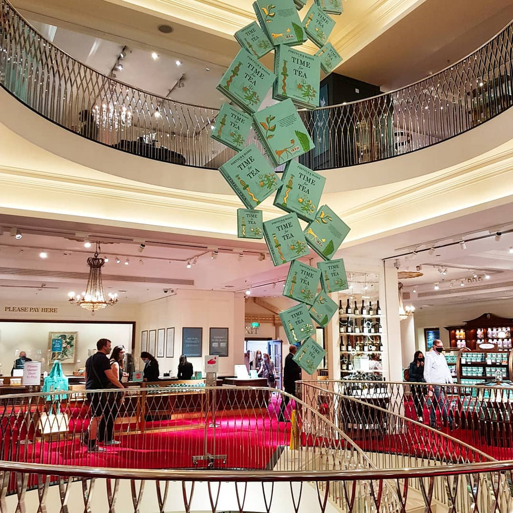 A cascade of pale green “Time for Tea” boxes suspended in the stairwell at Fortnum & Mason, with the red-carpeted stairs and shop floor below.