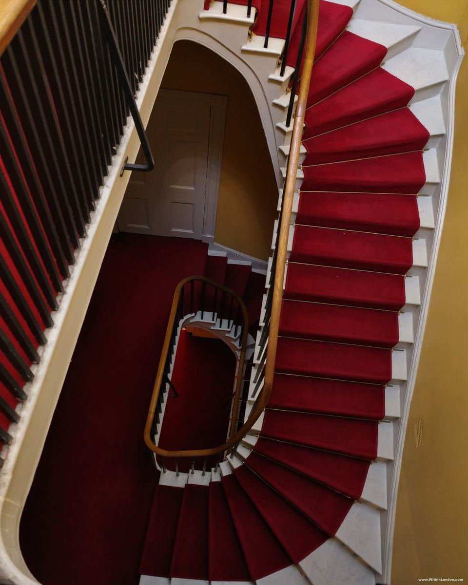 Looking down a curving palace staircase with a red carpet and a dark wooden handrail