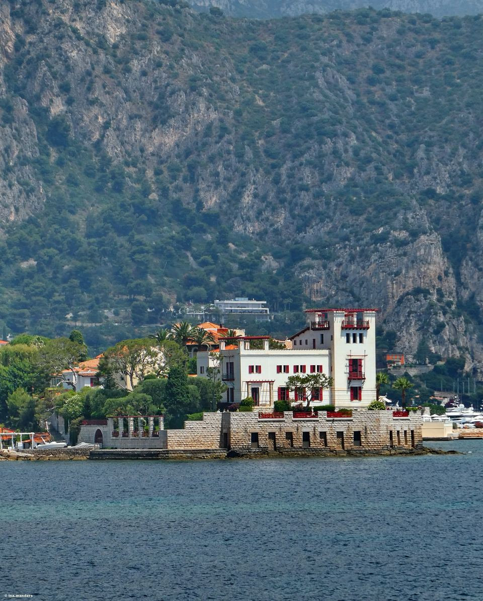 A white villa with a square tower and red shutters on a stone point at the water's edge, below steep wooded cliffs