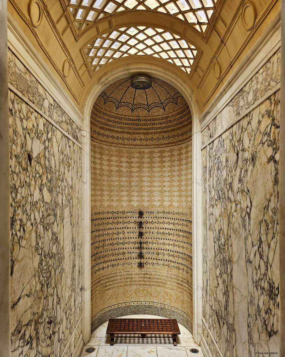 A tall arched shower niche lined with gold and black mosaic and marble walls, Greek lettering beside the fittings and a wooden slatted bench at the base