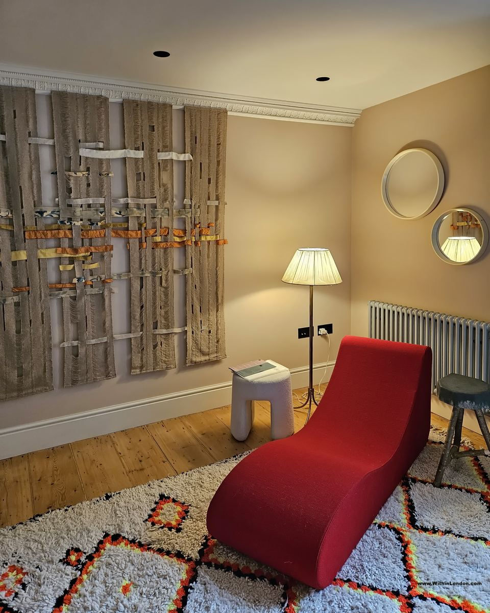 A cream bedroom with a curved red chaise longue on a red and black Berber rug, round mirrors on the wall and a screen of slim wooden rods threaded with orange cord