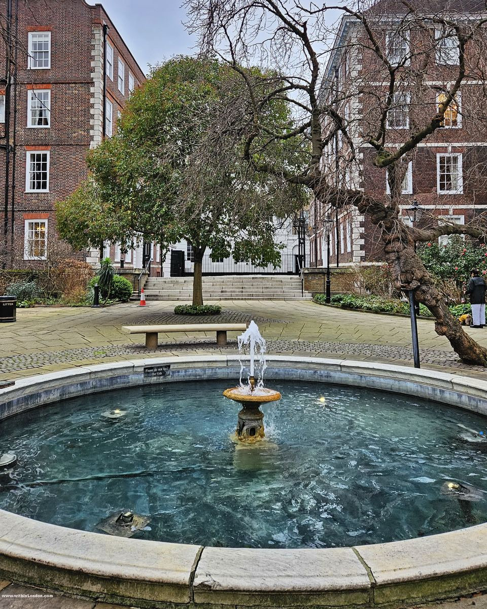 Round stone fountain basin with a small water jet, bare trees and red-brick chambers behind