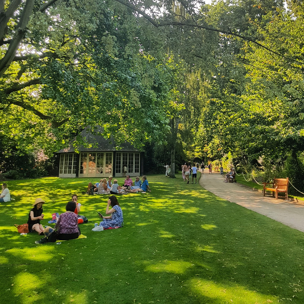 Visitors sitting on the lawn beneath large trees near a small garden pavilion in Buckingham Palace Gardens
