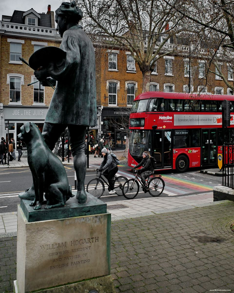 The bronze statue of William Hogarth and his pug on Chiswick High Road, with a red double decker bus and two cyclists passing behind it