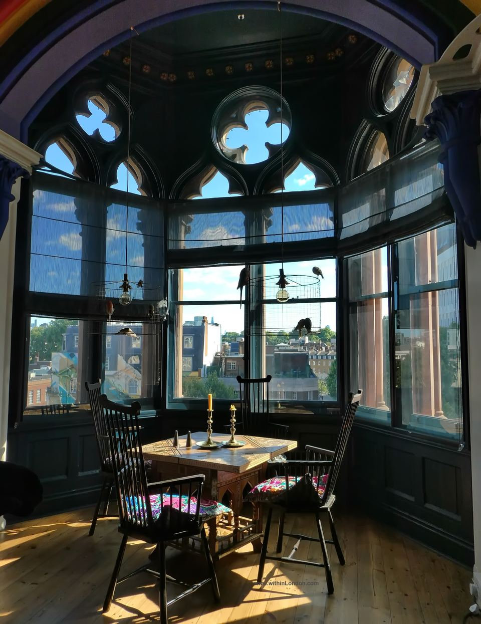 Wooden table with candlesticks and spindle-back chairs in a Gothic bay window, sunlight falling across the floorboards