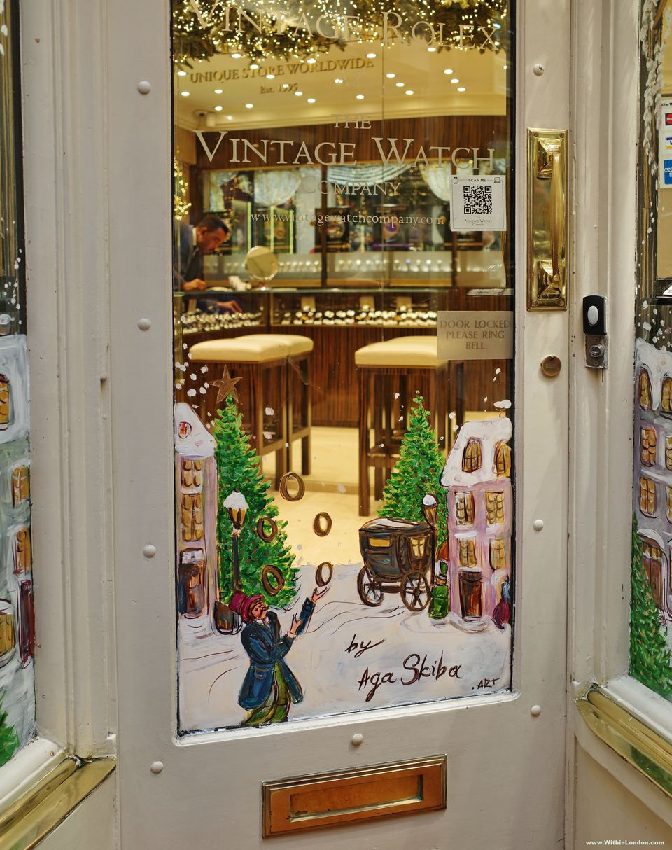 A shop door with a painted winter scene of a figure throwing gold rings beside a horse-drawn carriage and lit Christmas trees, signed Aga Skiba, with a watch shop's counter behind the glass