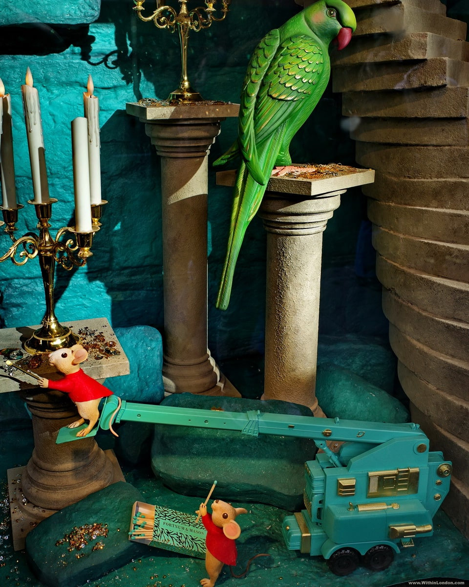 A green parrot on a stone column in a Fortnum & Mason window, with two mice in red coats and a turquoise toy crane below