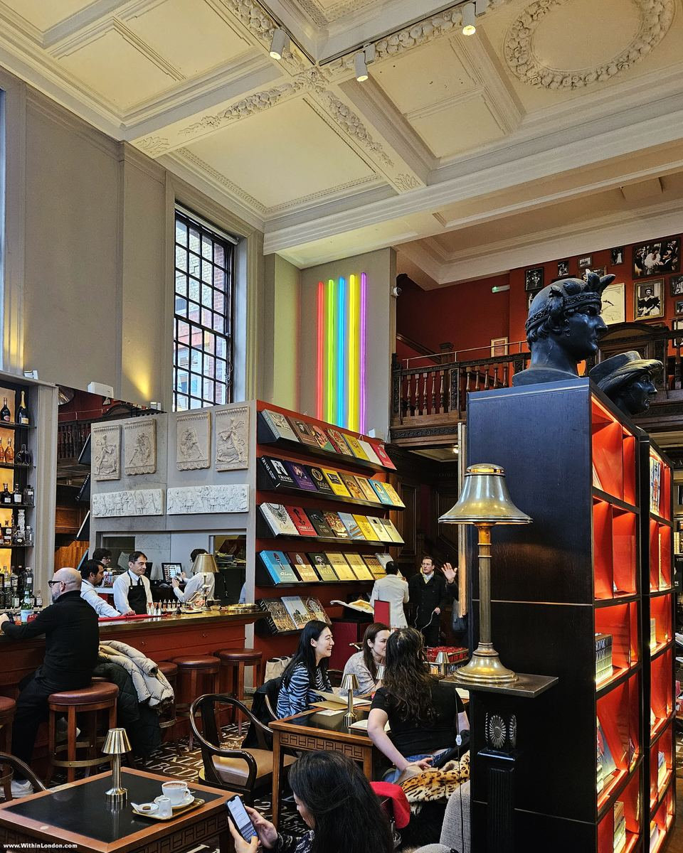 Bar with stools and people at small tables, a red-walled gallery above, a rainbow light strip on the wall and a dark bust on a bookcase lit red inside