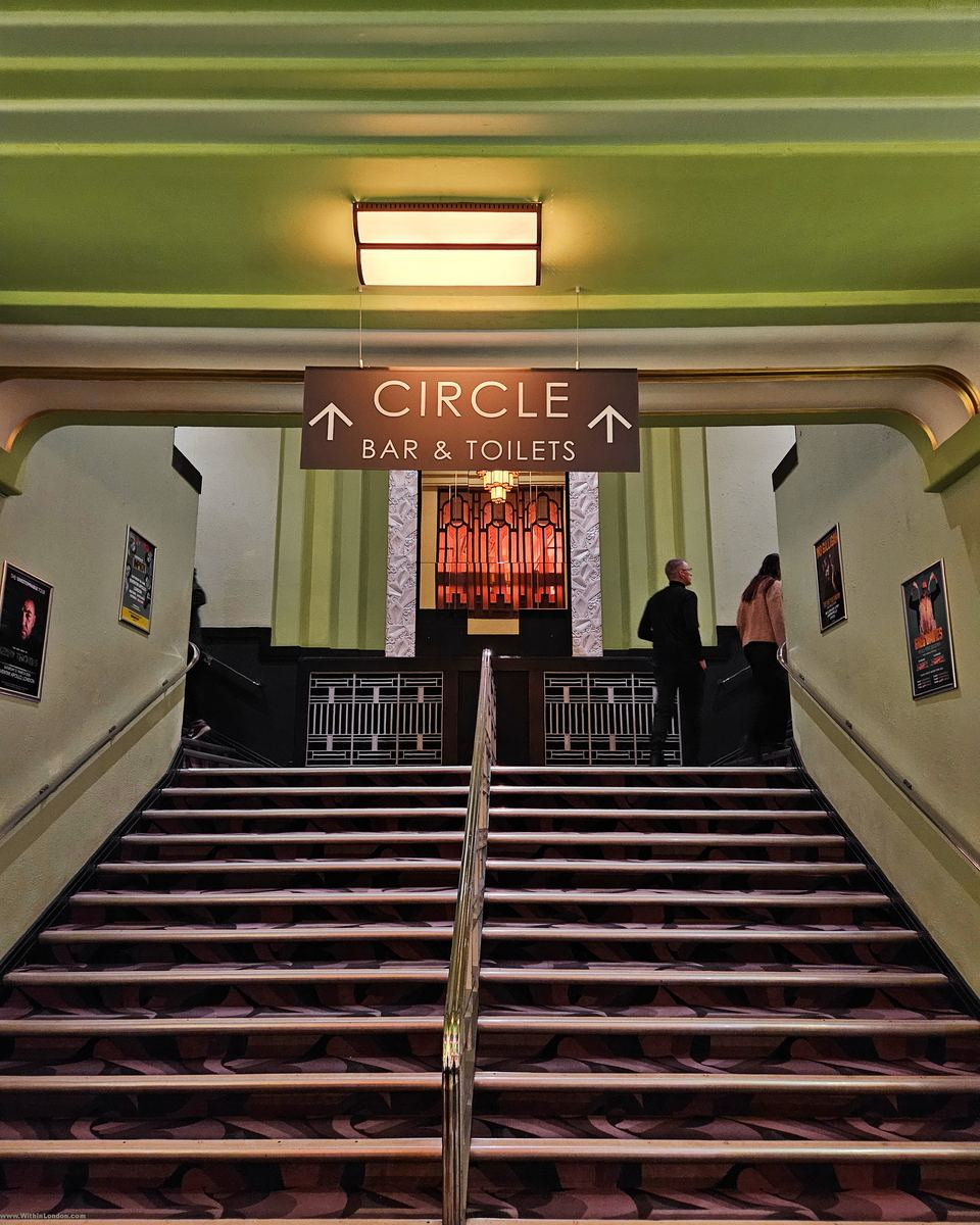 A wide Art Deco foyer staircase rising towards a lit Circle Bar sign
