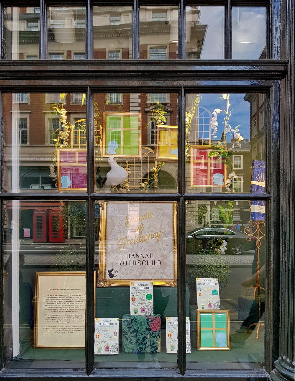 A Hatchards window display for a Hannah Rothschild novel, with painted model house fronts, a white bird ornament and books propped on a green shelf, seen from Piccadilly