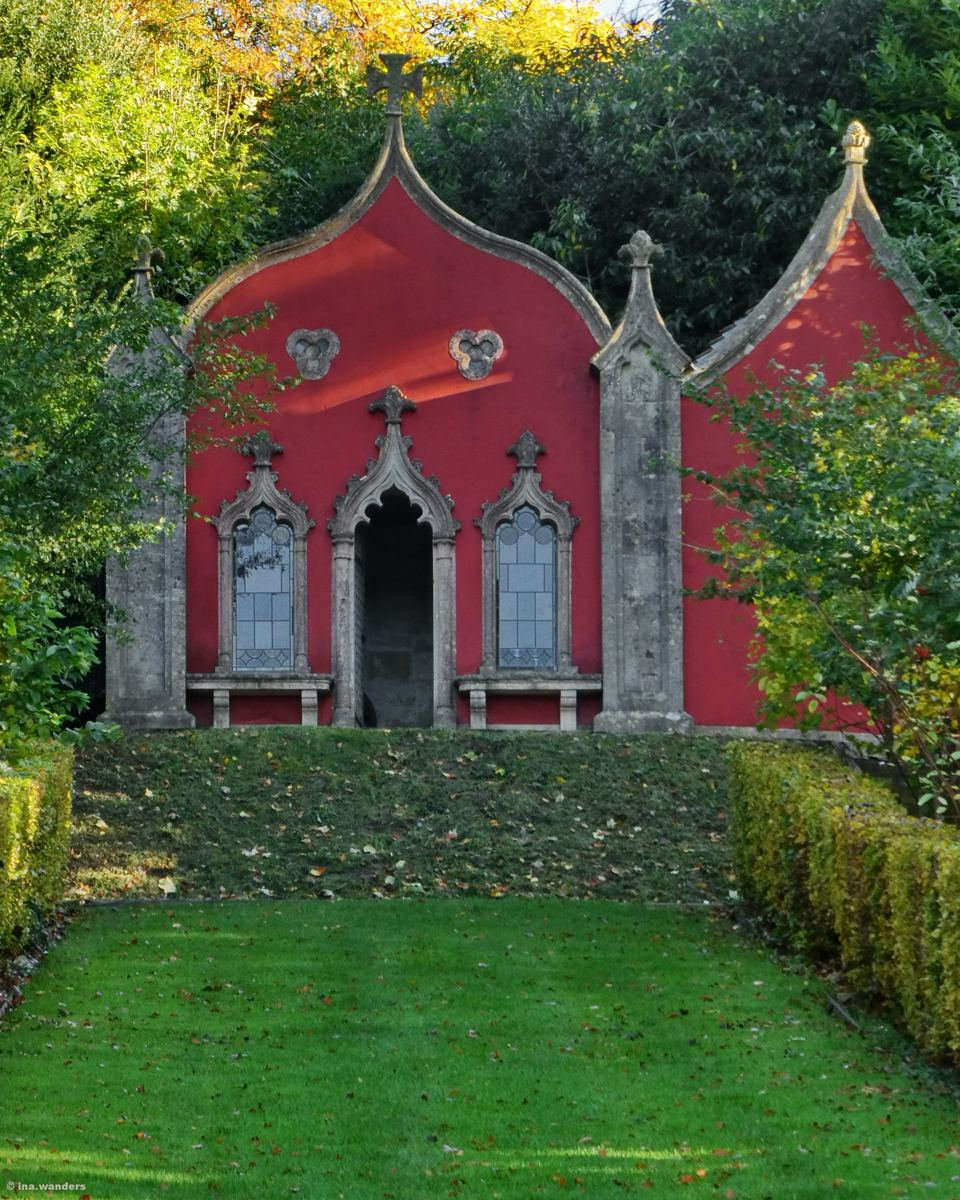 Red-painted Gothic building with curved gables and stone-framed windows behind a lawn and clipped hedges
