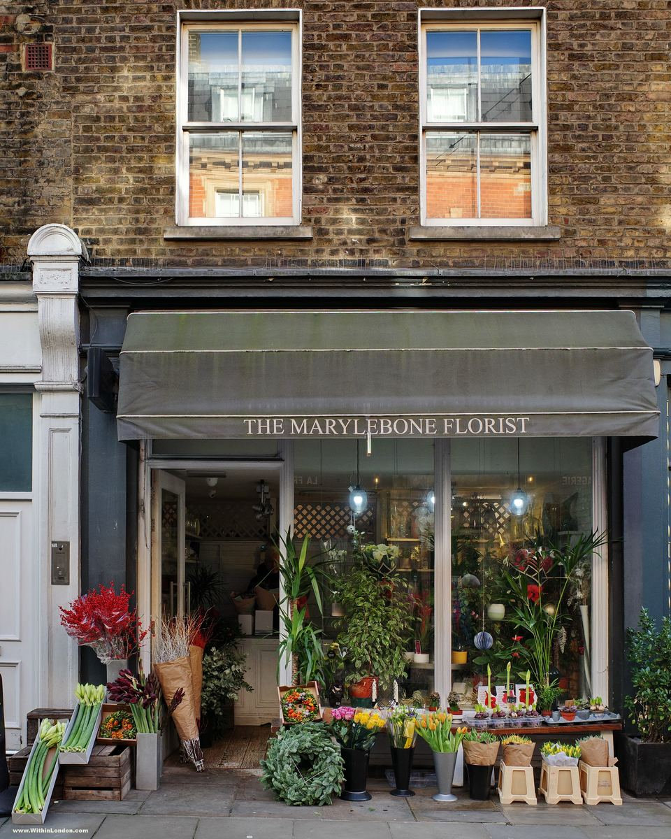 Buckets of flowers and small wreaths on the pavement outside The Marylebone Florist, under a grey awning
