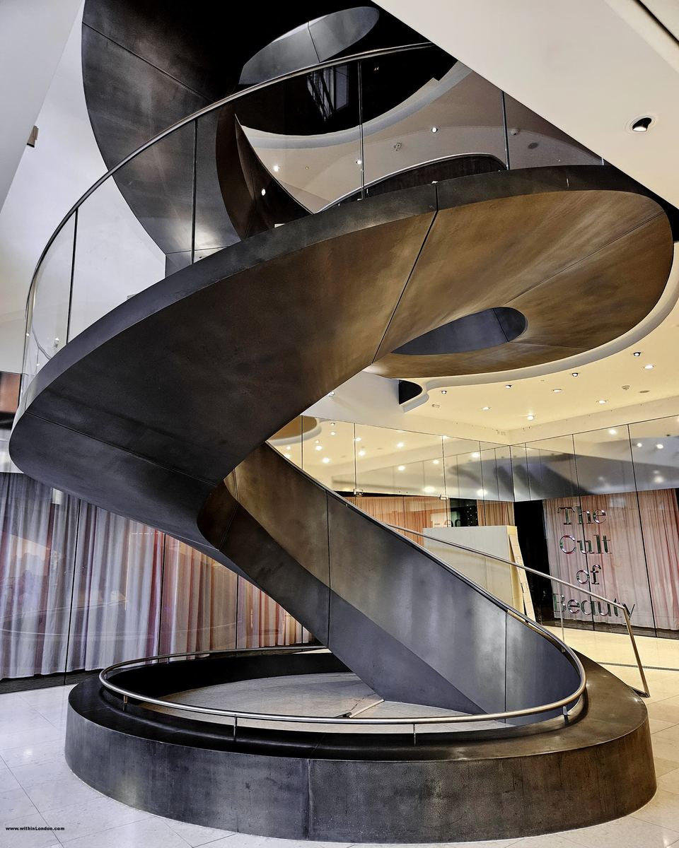A dark steel spiral staircase with pale stone steps coiling through a museum