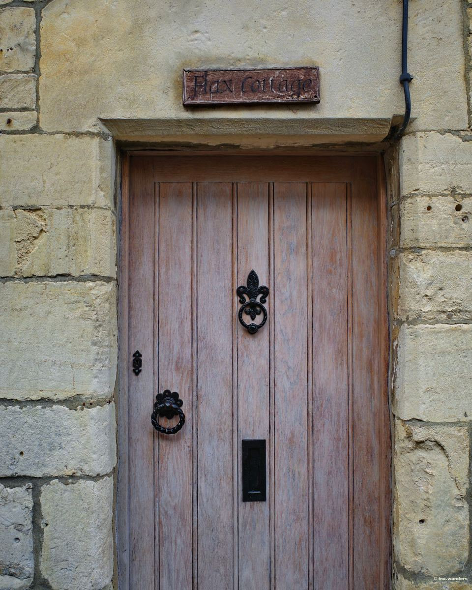 Weathered wooden door with an iron fleur-de-lis and ring handle under a carved name board in honey-coloured stone