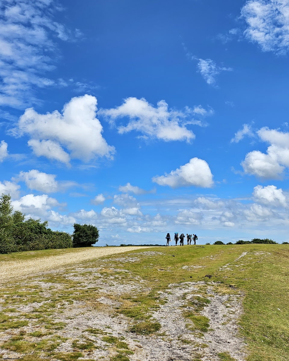 Walkers on a sandy track across open heath under a wide blue sky with white clouds